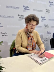 Eine Frau mit kurzen braunen Haaren und Brille sitzt an einem Tisch und schreibt mit einem Stift in ein Buch. Sie trägt einen beigen Pullover sowie einen bunt gemusterten Schal. Im Hintergrund ist eine Wand mit den wiederholten Schriftzügen „Buch Wien“ und „DER STANDARD“ zu sehen.