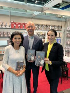 Drei Personen stehen lächelnd nebeneinander in einer Buchhandlung und halten Exemplare desselben Buchs in den Händen. Die Frau links trägt ein helles Kleid, der Mann in der Mitte trägt ein blaues Sakko und die Frau rechts trägt ein dunkles Sakko über einem gelben Oberteil. Im Hintergrund befinden sich Regale voller Bücher.