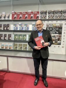 Ein Mann mit Brille steht lächelnd vor einem Bücherregal und hält ein rotes Buch in den Händen. Er trägt ein blaues Hemd, ein dunkles Sakko und eine dunkle Hose. Im Hintergrund sind Regale mit Büchern zu sehen.