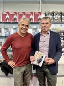 Zwei Männer stehen lächelnd nebeneinander vor einem Bücherregal. Der Mann links hat graues Haar und trägt einen rothen Langarmpullover. Der Mann rechts hat kurze braune Haare, trägt ein helles Hemd unter einem dunklen Sakko und hält ein Buch in der Hand, auf dem die Jahreszahl „1924“ zu sehen ist.