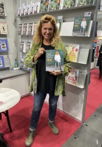 Eine Frau mit blonden Haaren steht lächelnd vor einem Bücherregal und hält ein quadratisches Buch vor sich. Sie trägt ein schwarzes Oberteil, eine gelb-grün gemusterte Überjacke und Jeans. Im Hintergrund sind Regale mit Büchern zu sehen.