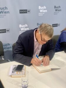 Ein Mann mit ergrauendem Haar und Brille sitzt vorneüber gebeugt an einem Tisch und signiert ein aufgeschlagenes Buch mit einem blauen Stift. Er trägt ein rosa Hemd und ein dunkles Sakko. Auf dem Tisch liegt zudem ein Smartphone. Im Hintergrund ist eine graue Wand mit dem Schriftzug „Buch Wien“ erkennbar.
