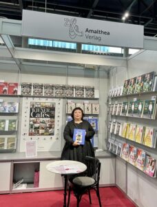 Eine Frau in schwarzer Kleidung steht in einem Messestand und hält ein blaues Buch in den Händen. Im Hintergrund sind Regale mit Büchern sowie ein Plakat mit der Aufschrift „Wiener Cafés“ zu sehen.