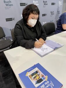 Eine Frau mit dunklen Haaren und weißer FFP2-Maske sitzt an einem Tisch und schreibt mit einem Stift in ein Buch. Sie trägt dunkle Kleidung. Im Hintergrund ist eine Messewand mit den Schriftzügen „Buch Wien“ und „Radio Wien“ zu sehen.