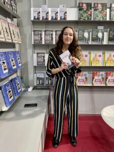 Eine junge Frau mit langen, dunkelbraunen Wellenhaaren steht lächelnd vor einem Bücherregal und hält ein Buch in beiden Händen. Sie trägt einen gestreiften Jumpsuit in Dunkelgrün, Schwarz und Gelb/Beige. Im Hintergrund sind mehrere Regale mit Büchern zu sehen.