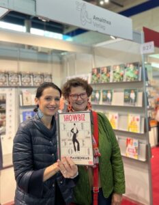 Zwei Frauen stehen nebeneinander in einer Buchhandlung oder auf einer Messe und halten gemeinsam ein Buch mit der Aufschrift „SHOWBIZ“ in die Kamera. Die links stehende Frau trägt eine dunkelblaue Steppjacke und hat hochstecktes dunkles Haar. Die rechts stehende Frau trägt eine grüne Jacke, einen roten Schal sowie eine rote Brille. Im Hintergrund sind Regale mit Büchern zu sehen.