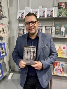 Ein Mann mit Brille steht lächelnd vor einem Bücherregal und hält ein Buch mit beiden Händen in die Kamera. Er trägt ein dunkles Hemd und ein graues Sakko. Im Hintergrund sind Regale mit mehreren Exemplaren desselben Buchs zu sehen.