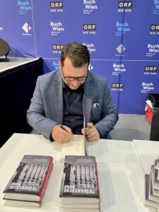 Ein Mann mit Brille sitzt an einem Tisch und signiert ein Buch mit einem Stift. Er trägt ein dunkles Hemd und ein graues Sakko. Auf dem Tisch liegen weitere Bücher. Im Hintergrund ist eine blaue Wand mit den Logos von „Buch Wien“ und „ORF“ zu sehen.